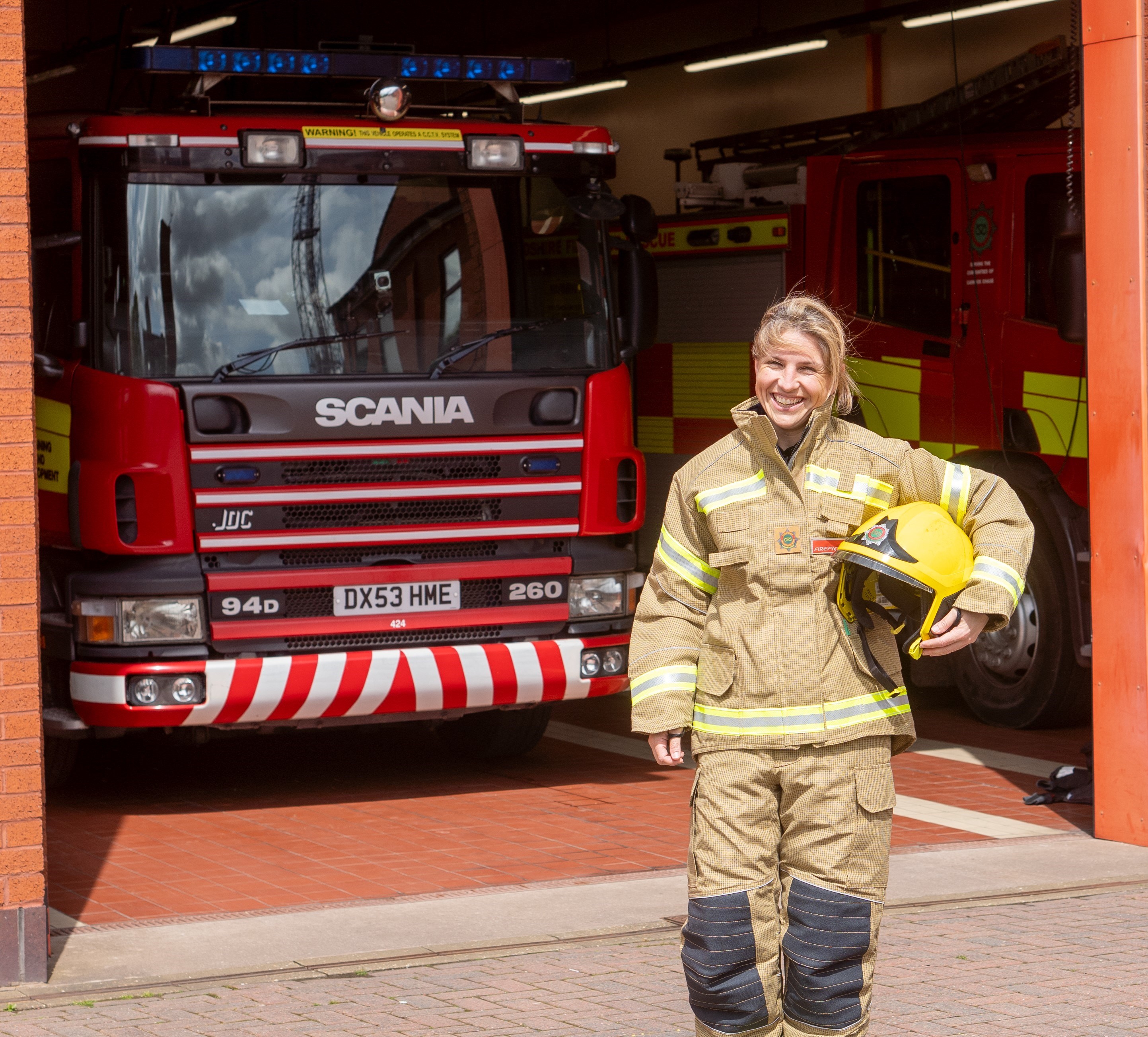 Female firefighter stood holding her helmet in front of an appliance bay with a fire engine parked inside