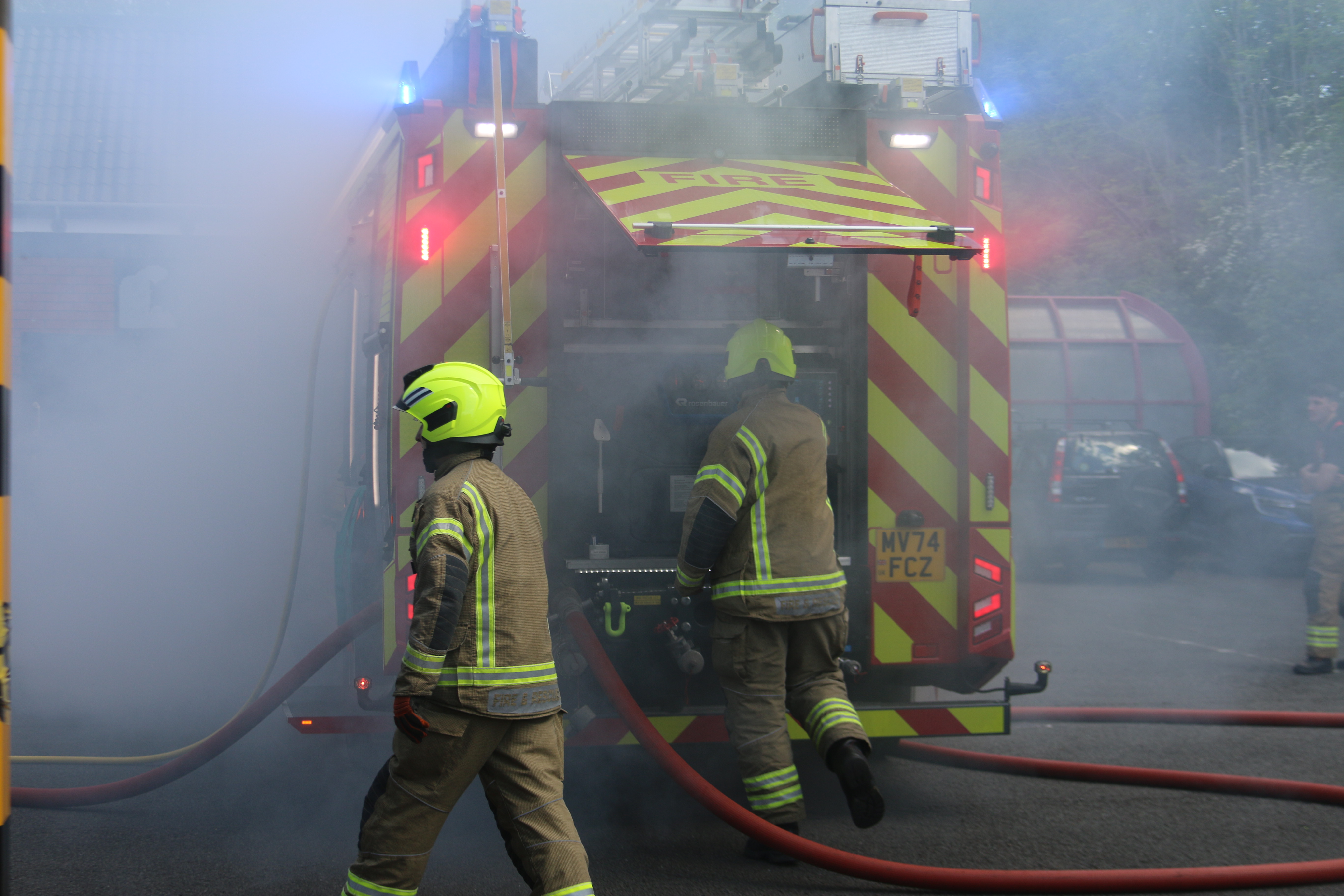 Two firefighters at the rear of a fire engine which is engulfed in smoke